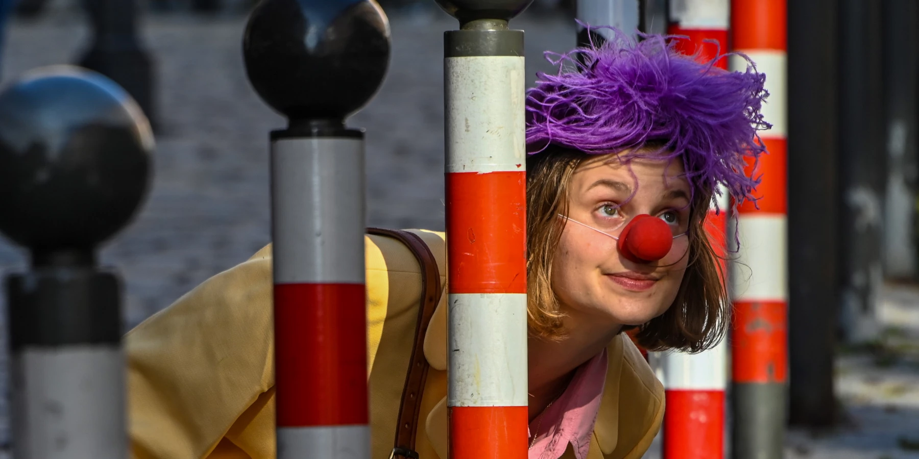 Eine Frau mit Clown-Nase und Lila Perücke blickt zwischen gestreiften Pollern hervor.