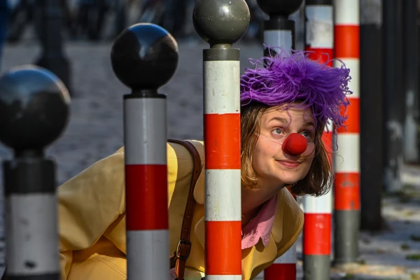 Eine Frau mit Clown-Nase und Lila Perücke blickt zwischen gestreiften Pollern hervor.