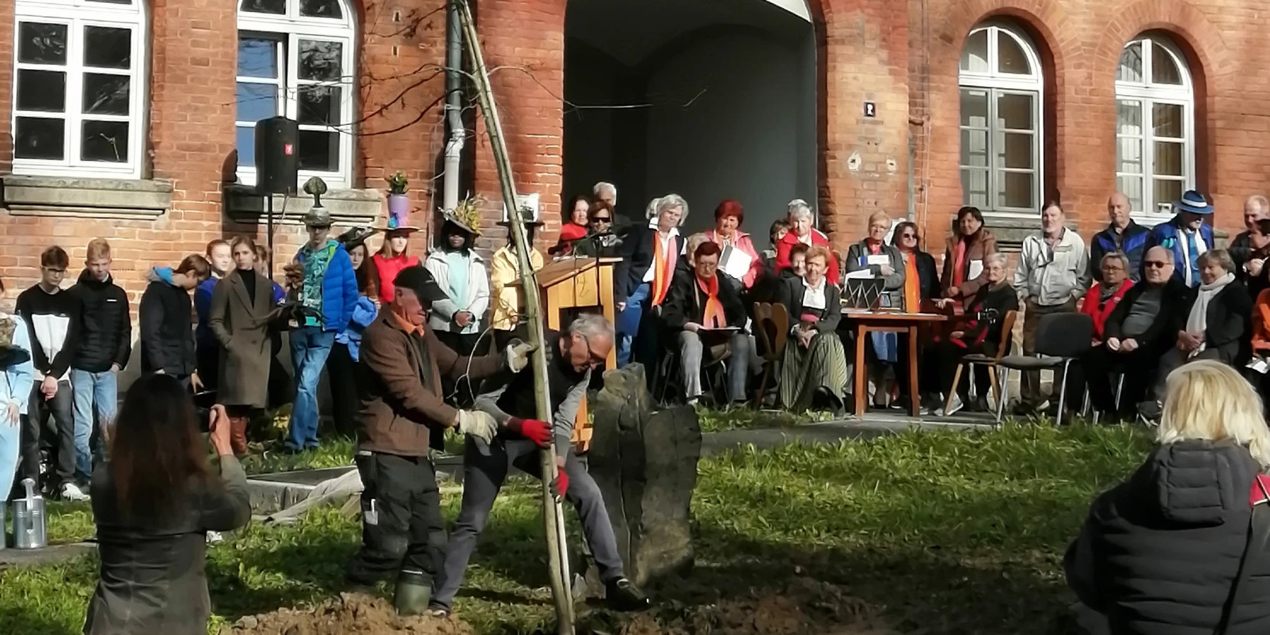 Vor einem roten Backsteinhaus wird eine Eiche gepflanzt, viele Menschen schauen zu