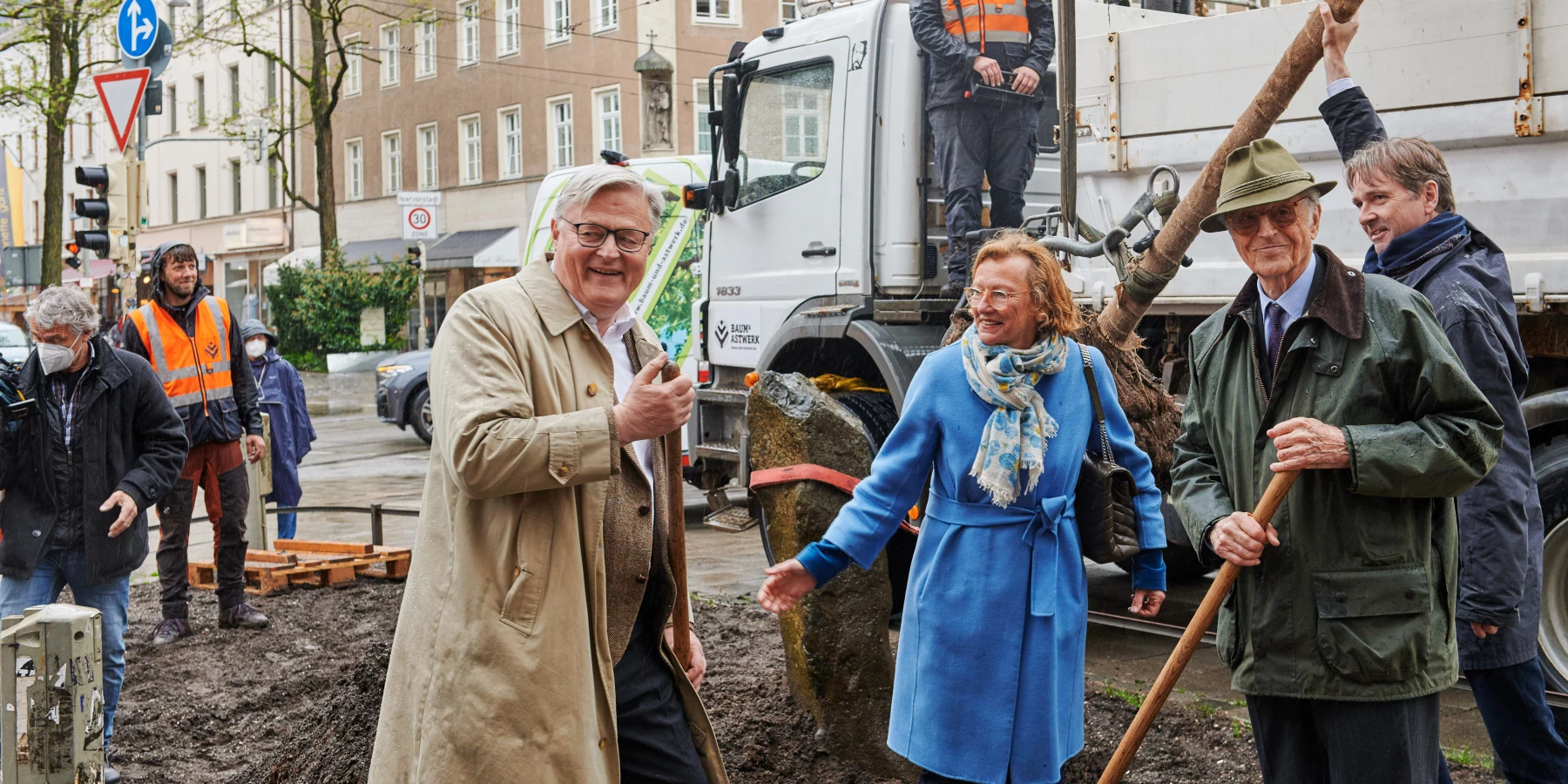 Drei Menschen im Vordergrund mit einer Schaufel. Im Hintergrund ein Bagger und Menschen in Arbeitskleidung