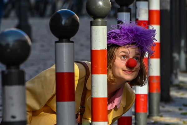 Eine Frau mit Clownsnase und lila Kopfschmuck steckt ihren Kopf durch eine Lücke von Pollern