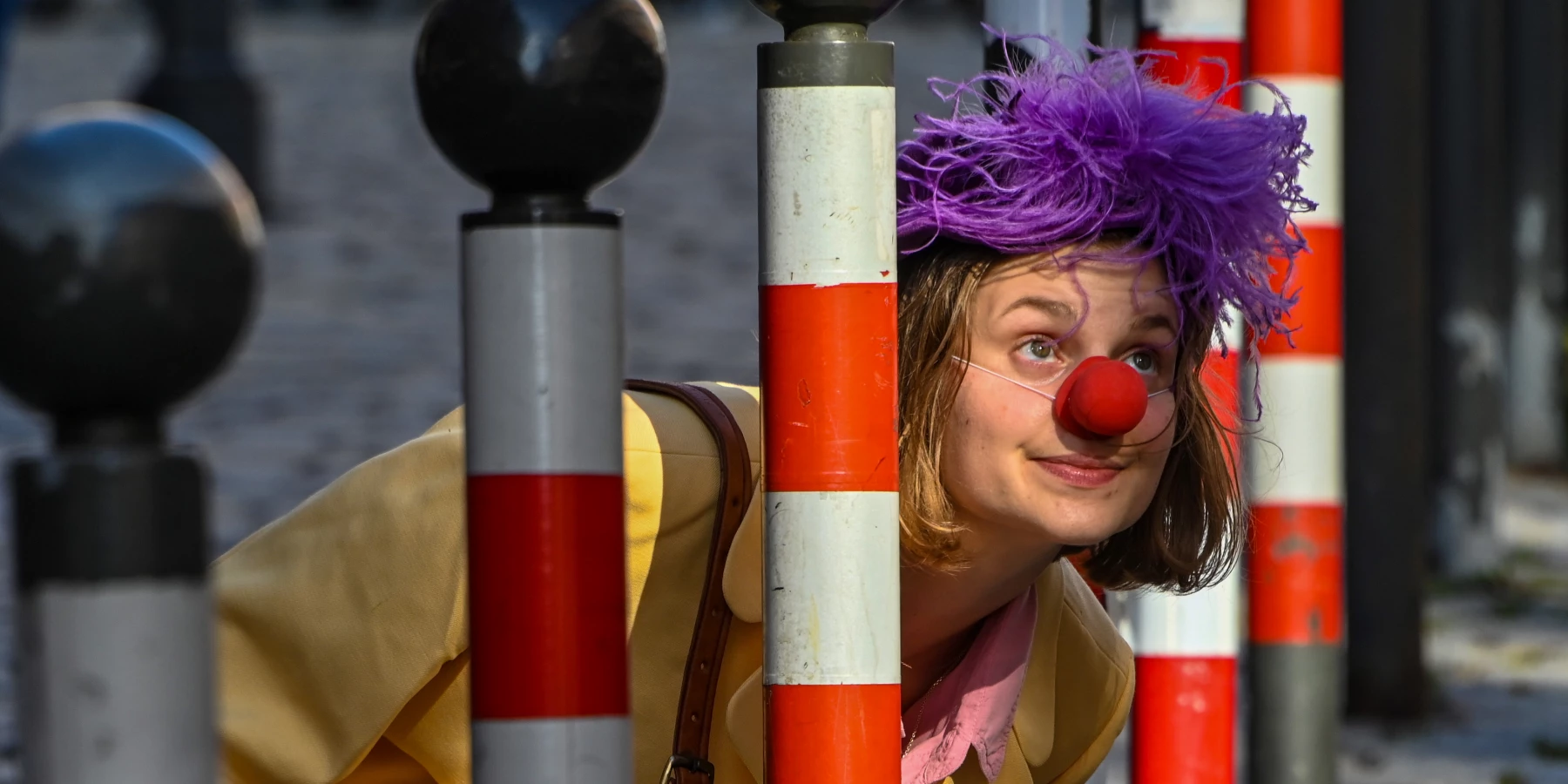 Eine Frau mit Clownsnase und lila Kopfschmuck steckt ihren Kopf durch eine Lücke von Pollern