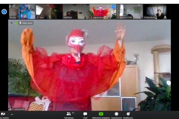 Gruppen Videocall Screenshot mit einer rot gekleideten Figur in der Mitte mit Schnabelmaske, die die Arme flügelartig ausbreitet