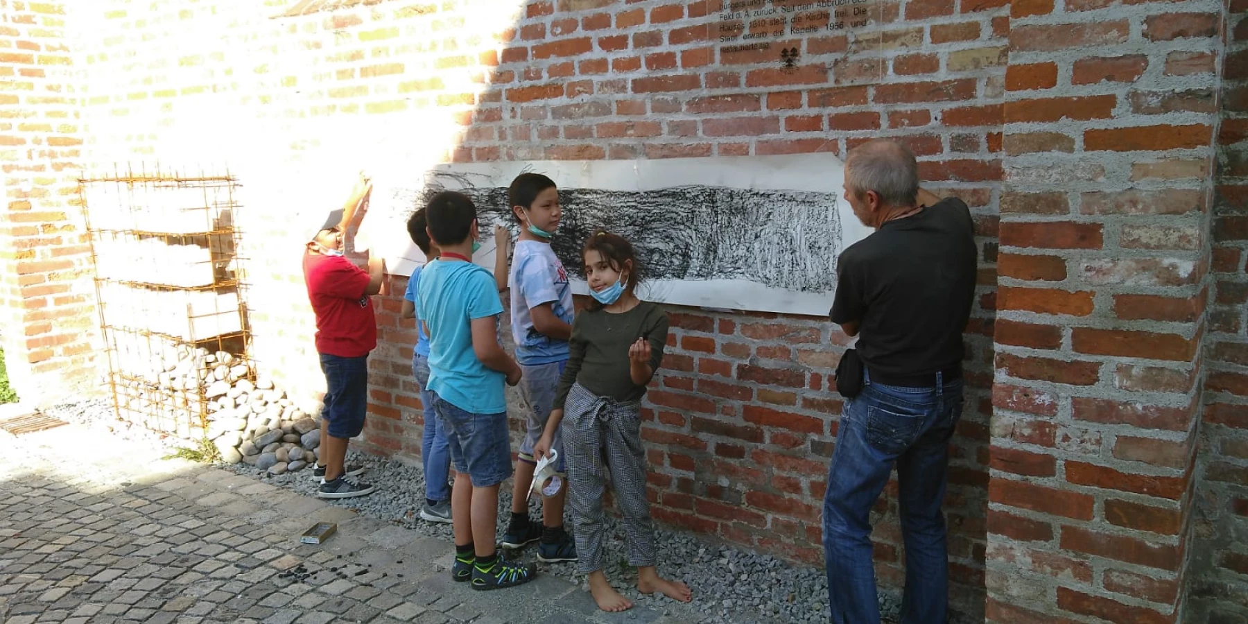 Kinder malen unter Anleitung eines Erwachsenen an einem langen Blatt Papier, welches an einer Außenwand mit Ziegelsteinen befestigt ist