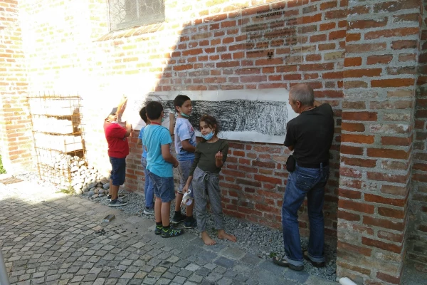Kinder malen unter Anleitung eines Erwachsenen an einem langen Blatt Papier, welches an einer Außenwand mit Ziegelsteinen befestigt ist
