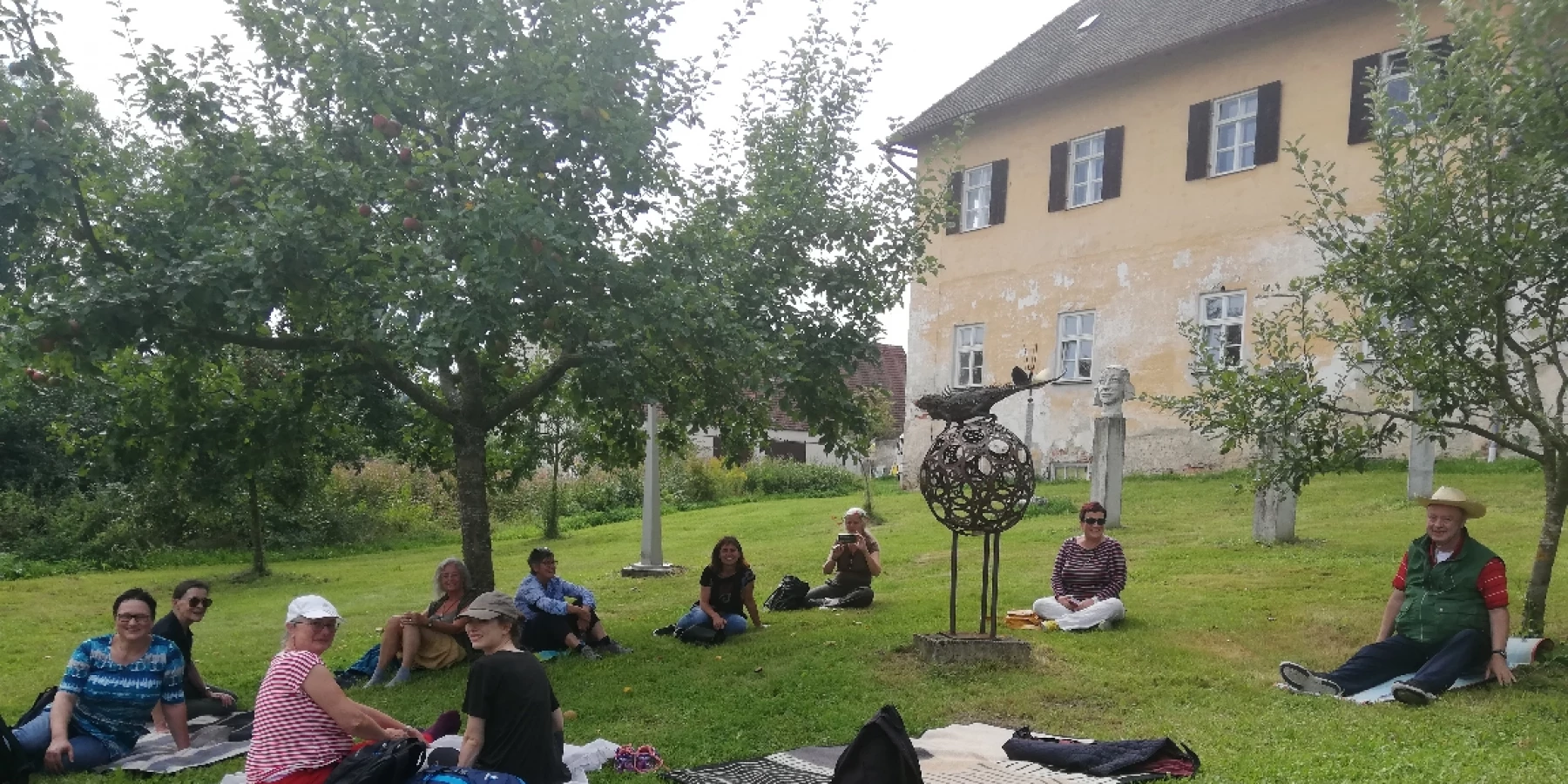 Die WorkshopteilnehmerInnen sitzen in einem Garten auf Decken, im Hintergrund ist ein altes Gebäude