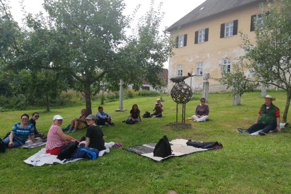 Die WorkshopteilnehmerInnen sitzen in einem Garten auf Decken, im Hintergrund ist ein altes Gebäude