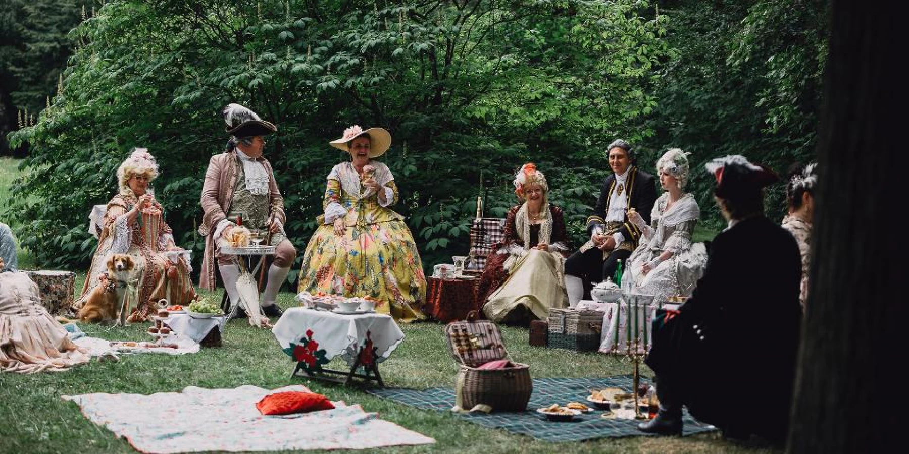 Damen und Herren in Rokokokostümen sitzen beim Picknick im Park