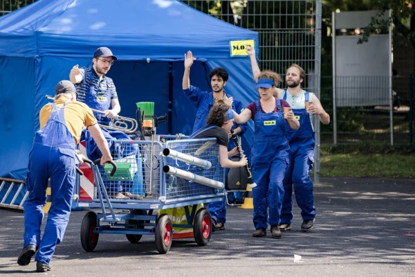 Ein Gruppe Menschen in blauen Overalls schiebt einen Wagen, manche gestikulieren