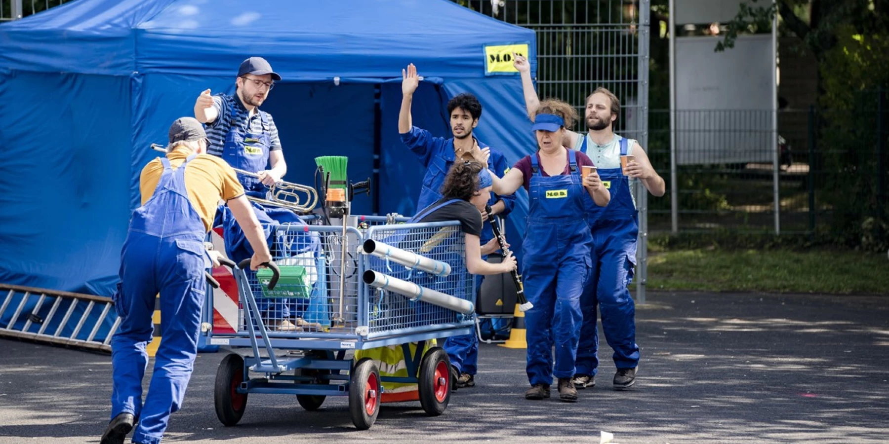 Ein Gruppe Menschen in blauen Overalls schiebt einen Wagen, manche gestikulieren