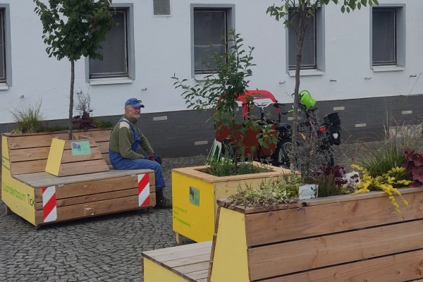 Ein Mann sitzt auf einer Holzbank, es stehen Holzkästen mit Bäumen daneben, sowie auf der Bank integriert