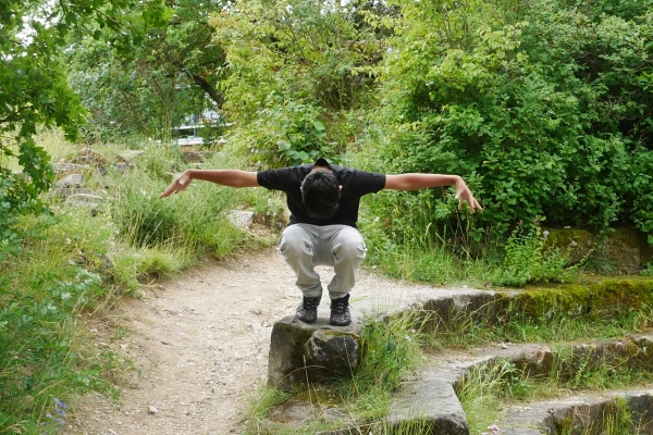 Auf einem Baumstamm geht ein Junge in die Hocke, beide Arme flügelartig ausgebreitet