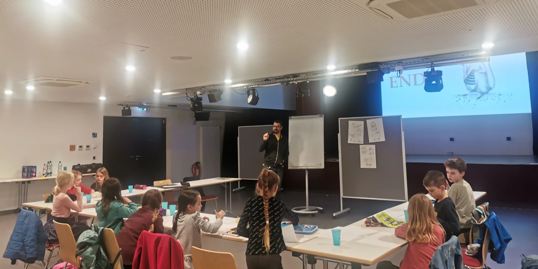 Kinder sitzen an Tischen, während ein Mann eine Präsentation hält