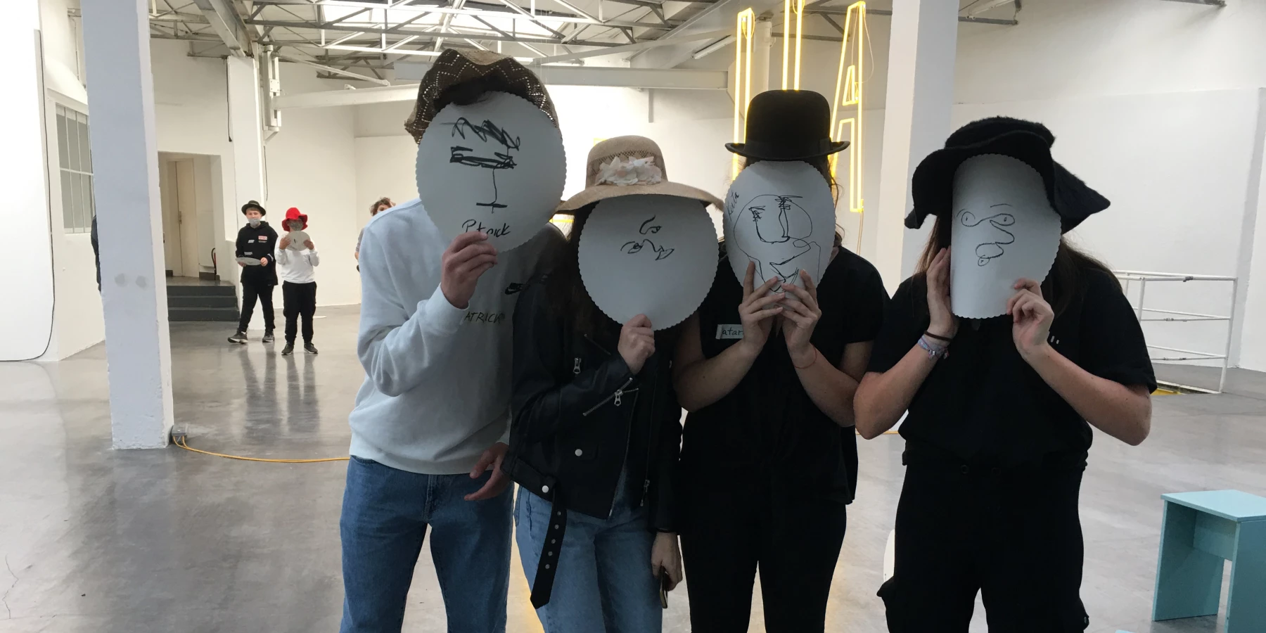 In einem Ausstellungsraum halten vier junge Menschen Gesichter aus weißen, bemalten Papptellern