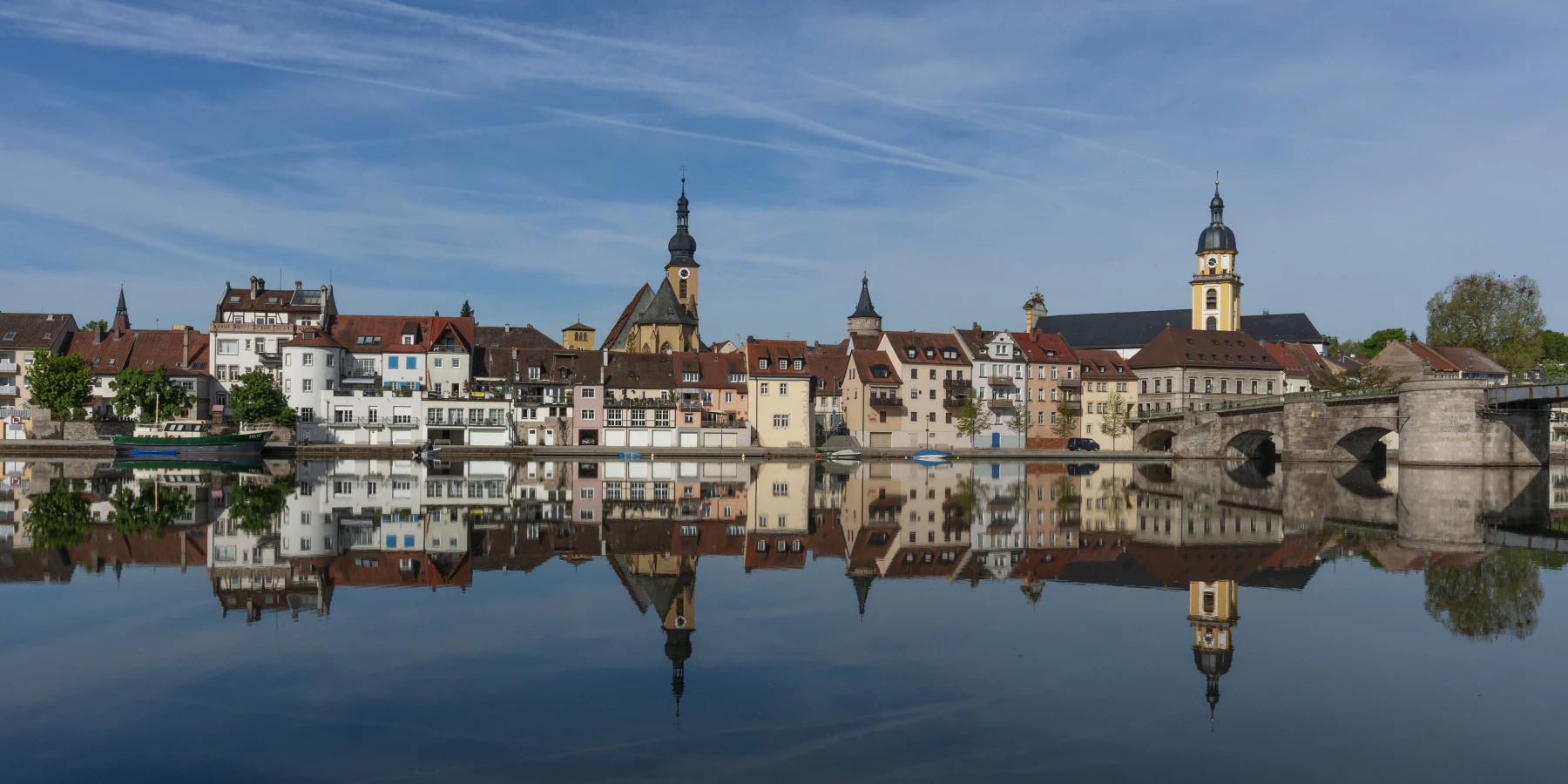 Eine Silhouette von Kitzingen mit Kirchen und rechts einer Brücke spiegelt sich im Wasser