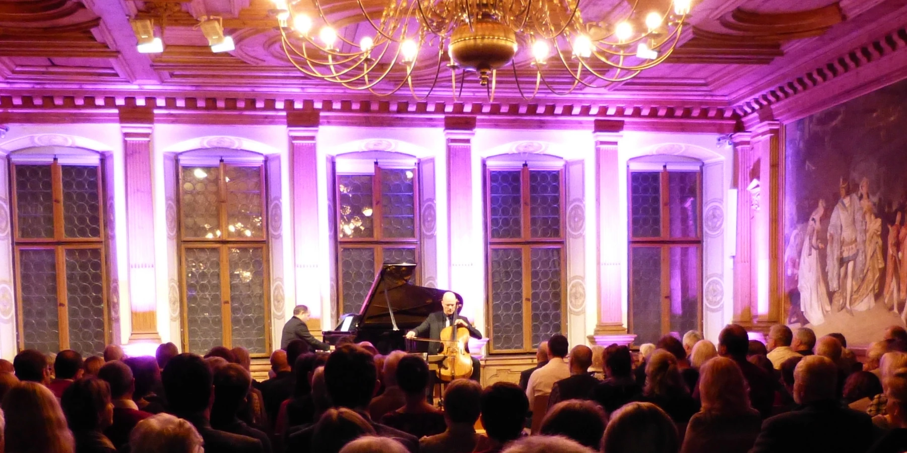 Ein Pianist und ein Cellist spielen vor Publikum in einem pink beleuchteten, opulenten-ausgestatteten Saal.