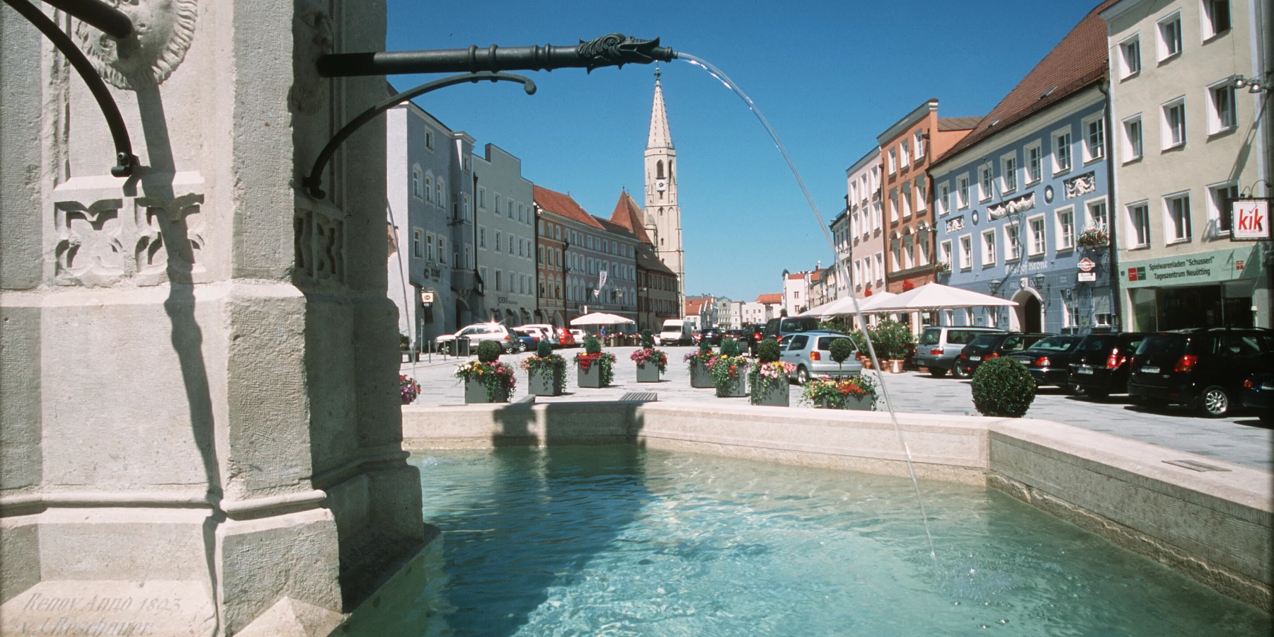 Ein alter Steinbrunnen im Vordergrund; Cafés, Häuser und Kirche im Hintergrund