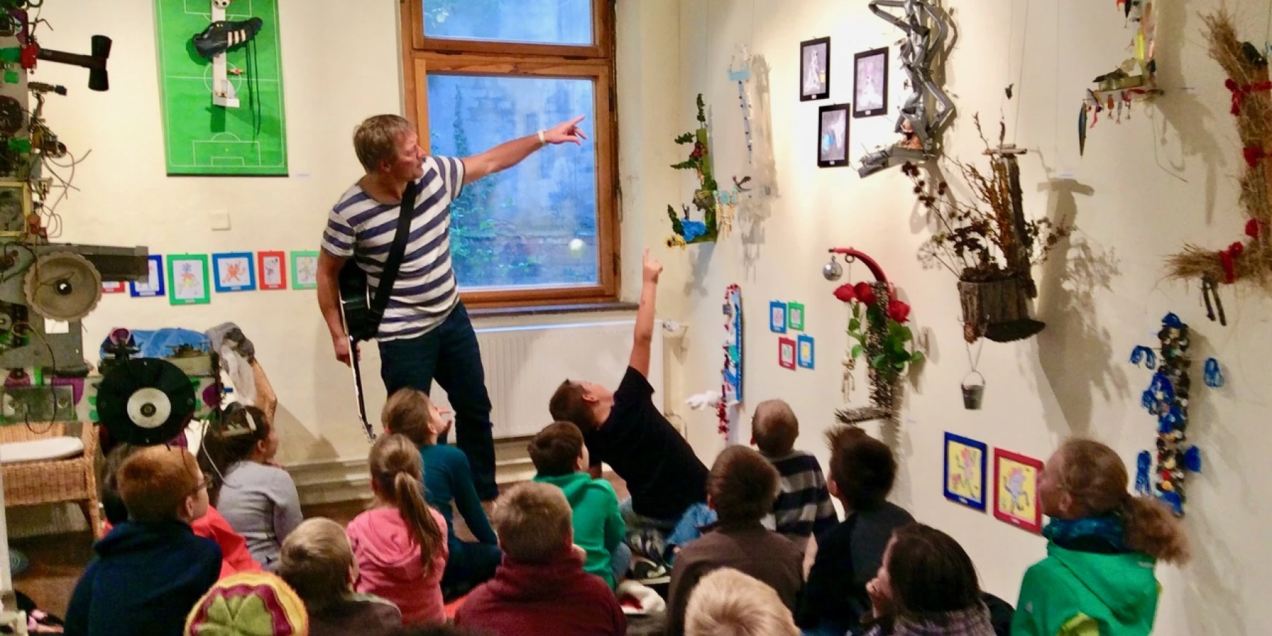 Ein Workshopleiter mit Gitarre zeigt vor einer Gruppe von sitzenden Kindern auf ein Objekt, das rechts an einer Wand hängt