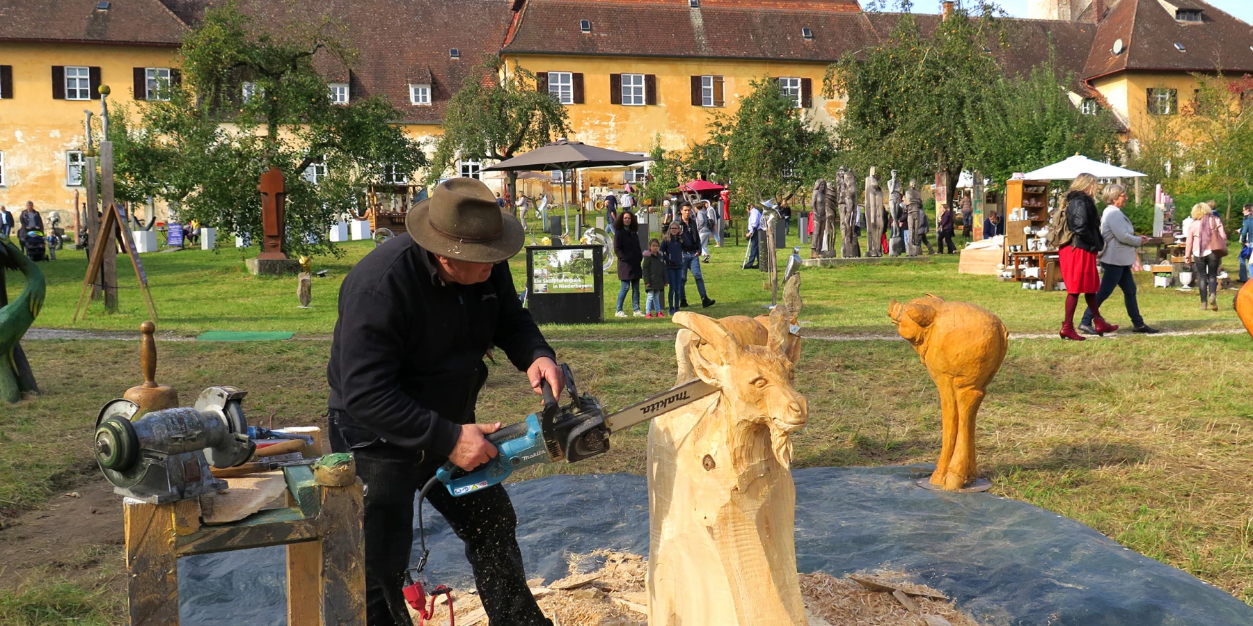 Ein Mann mit elektrischer Säge schnitzt einen Ziegenbock aus Holz, in einem Park mit einem gelben Gebäude im Hintergrund