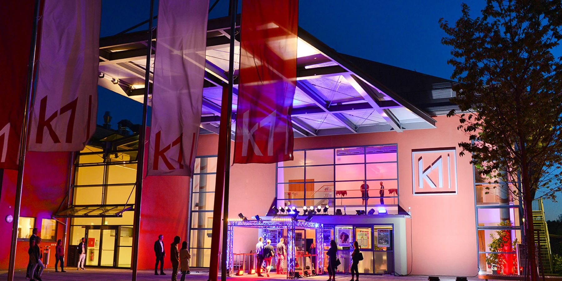 Bunt beleuchtete Außenansicht vom k1 Gebäude, Fahnen wehen draußen mit dem k1 Logo